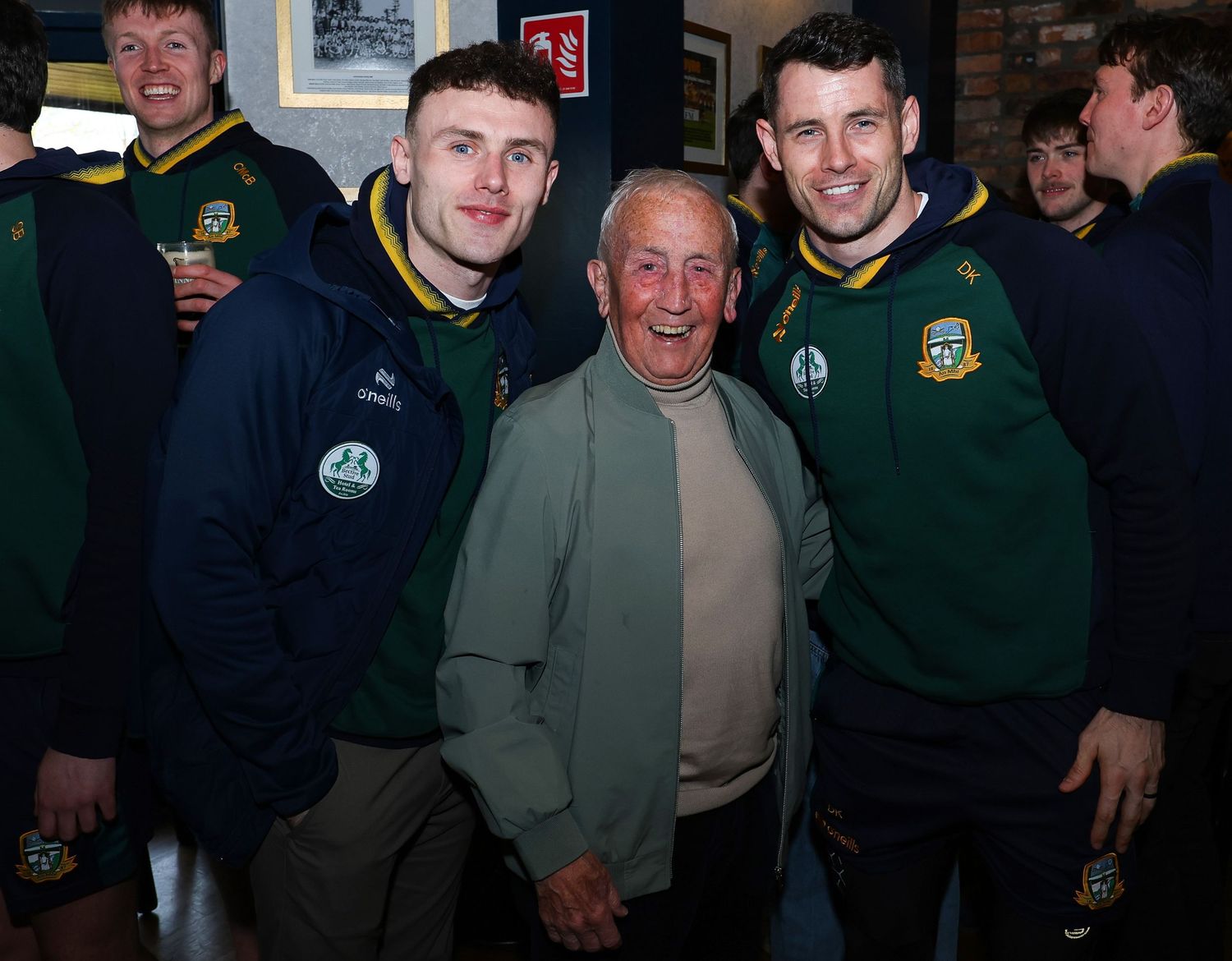 Three men in green and gold sports jackets pose together smiling inside a pub or club venue.