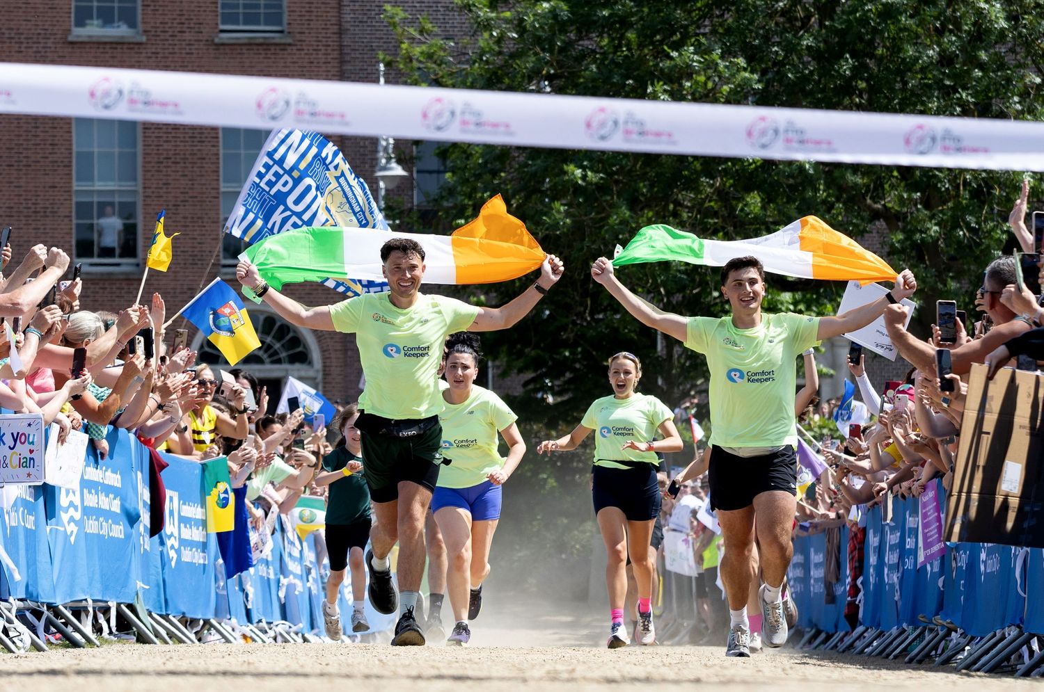 Runners carrying Irish flags cross the finish line at an outdoor race event cheered on by spectators.