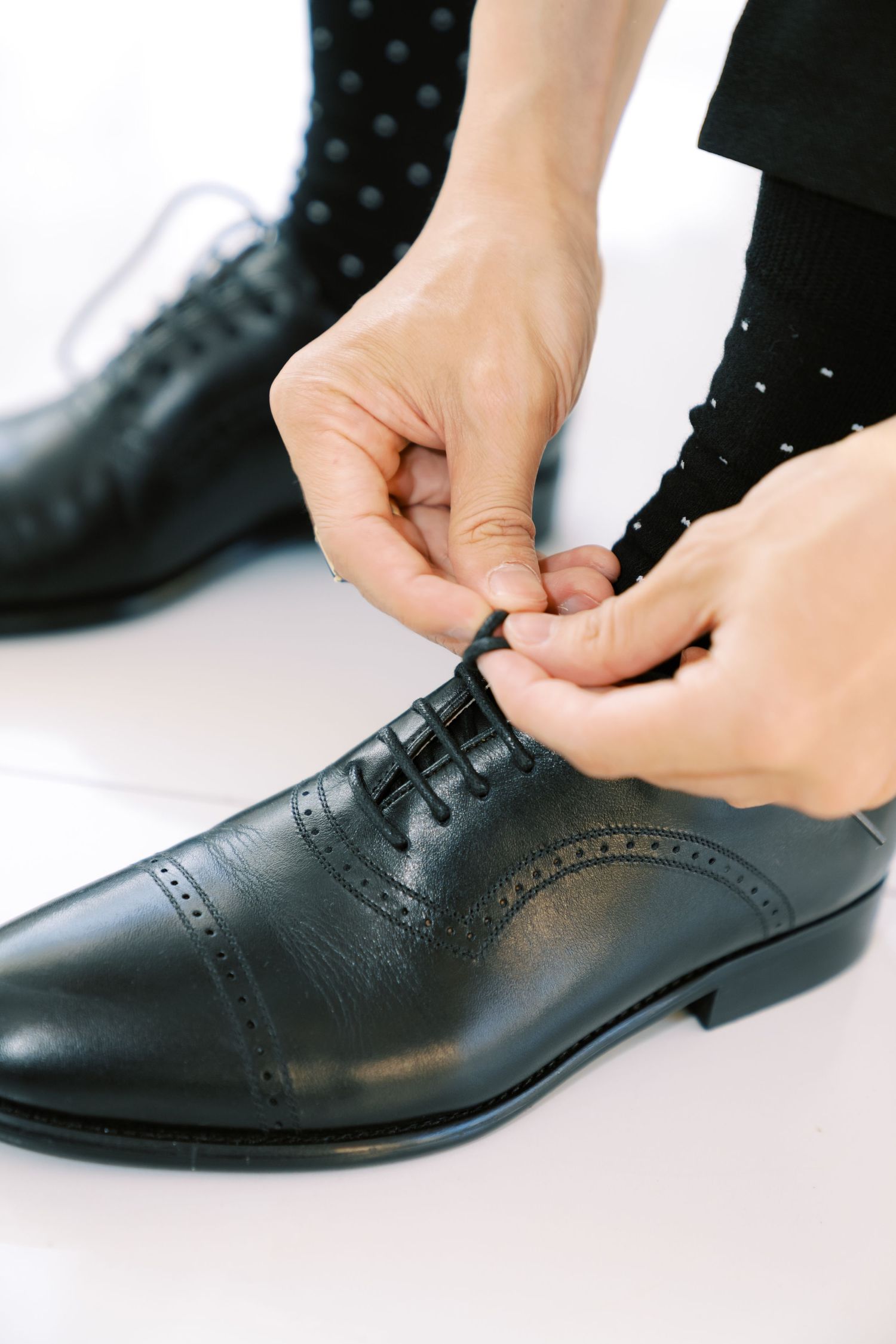 Person tying laces on a formal black leather oxford dress shoe with cap toe detailing.