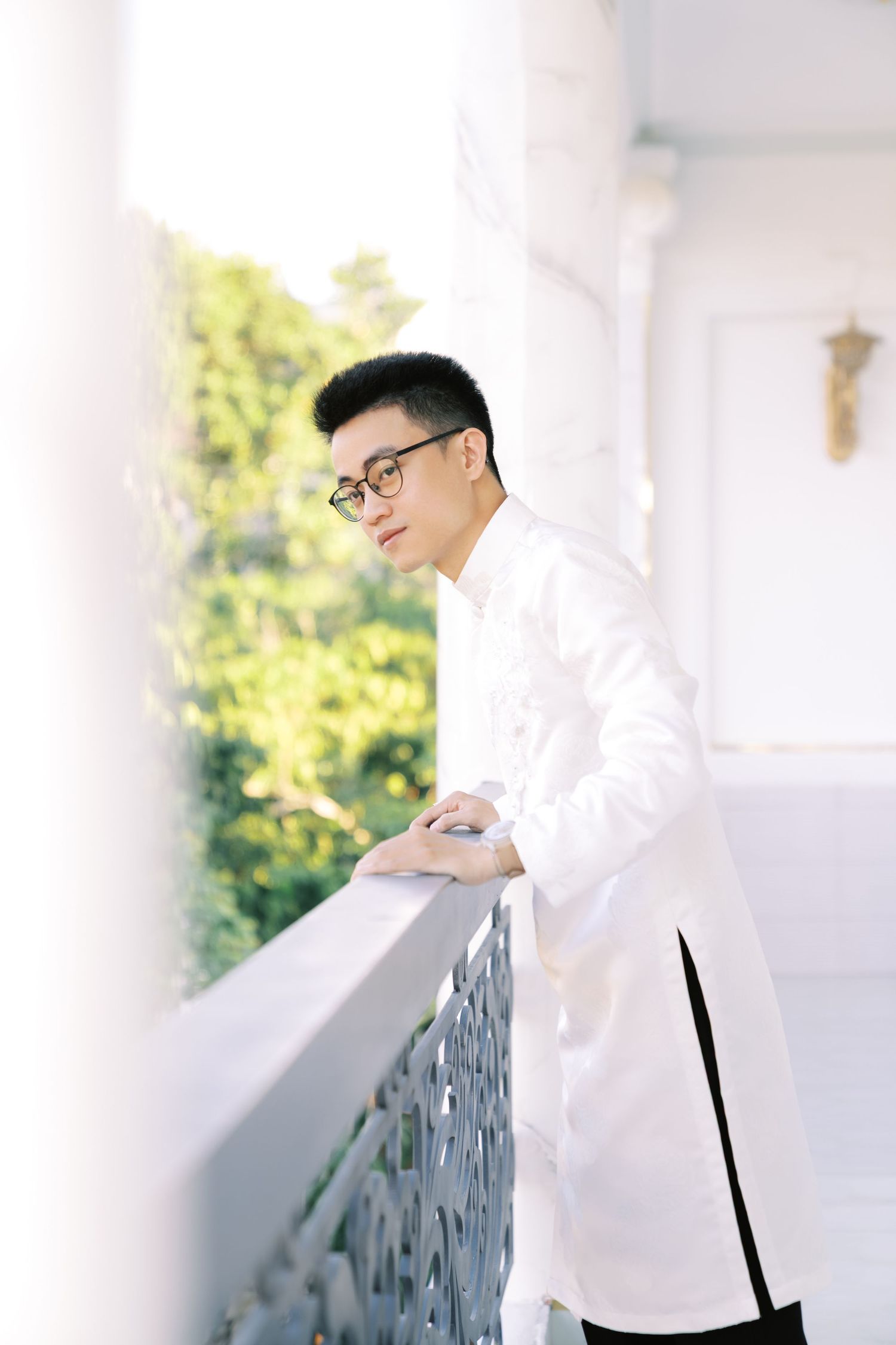 Person in glasses and white outfit leaning on balcony railing with greenery in background.