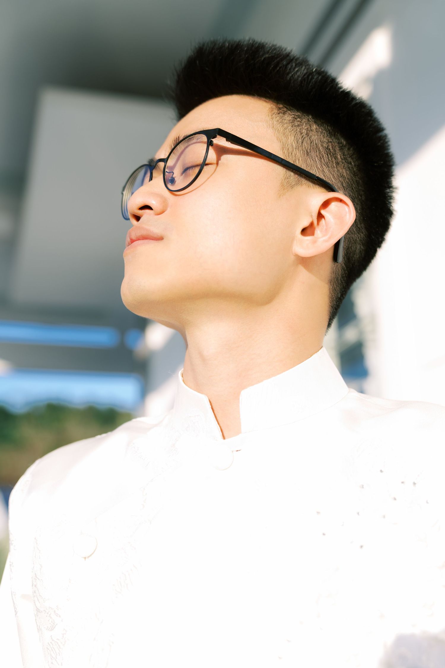 A young Asian person wearing glasses and a white shirt poses with eyes closed in natural sunlight.