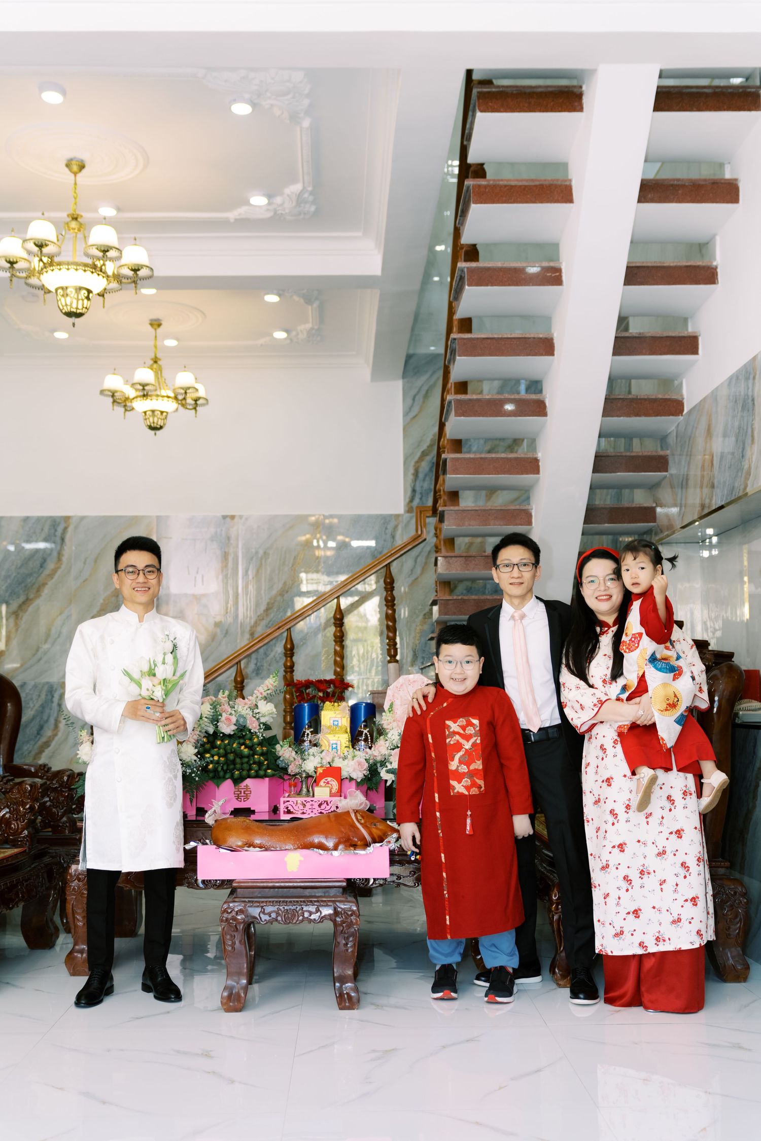 People in traditional Asian attire gather around decorative table with food display in elegant interior with chandeliers.
