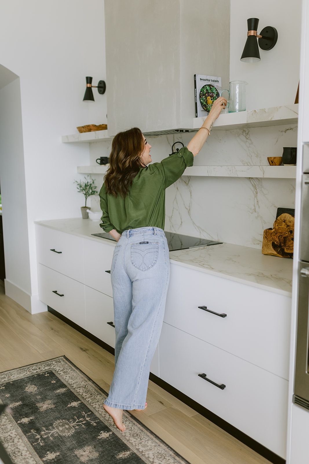 Person in green shirt and light jeans reaching up to white floating shelves in modern white kitchen with marble countertops.
