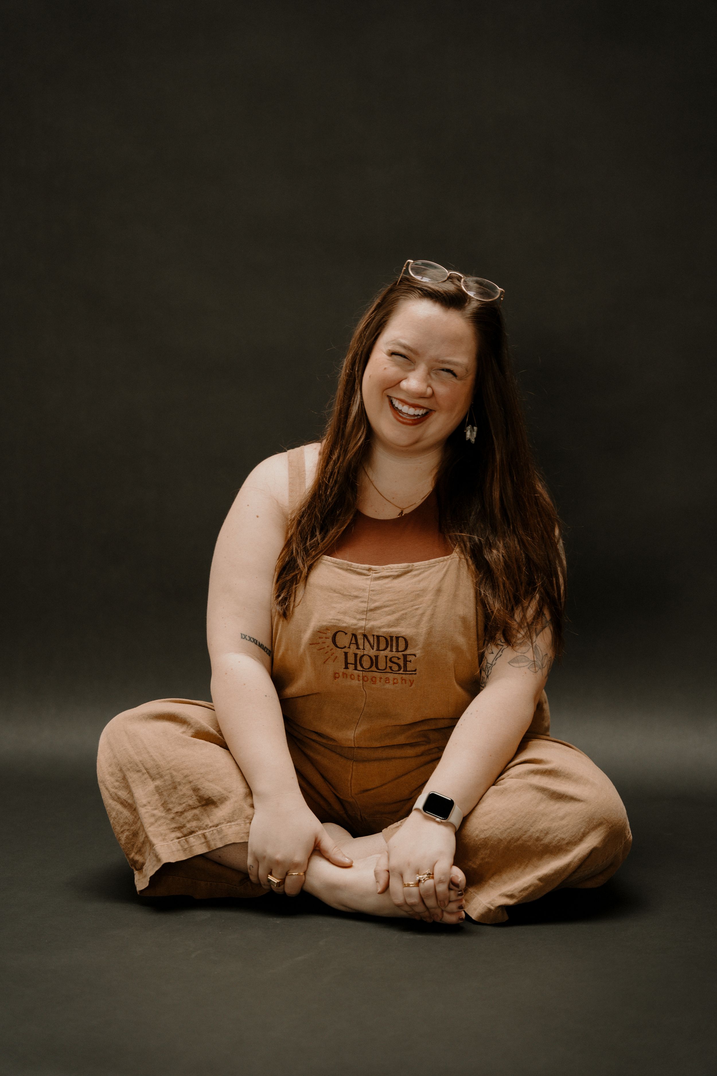 Person in beige overalls sits cross-legged on dark background, smiling joyfully while wearing sunglasses on head.