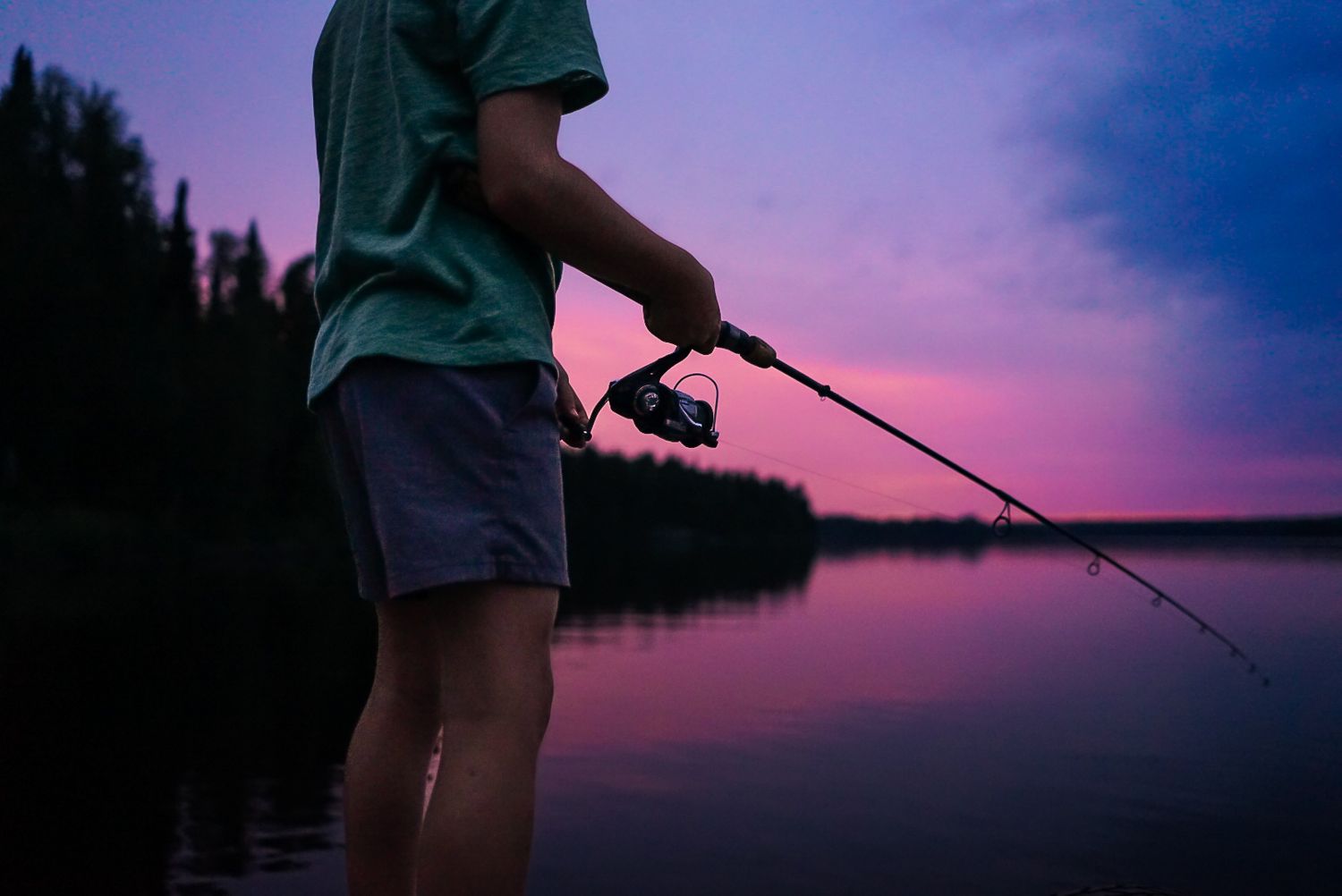 Silhouette of fishing rod against purple sunset sky over tranquil lake.