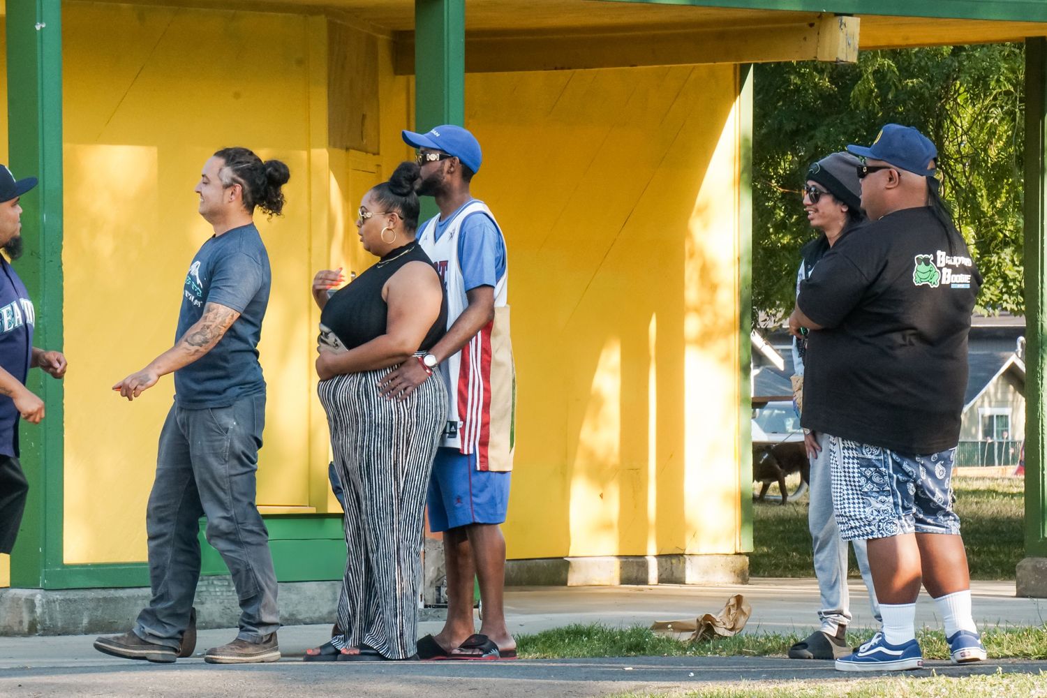 People wait in line at a yellow bus stop shelter on a sunny day.