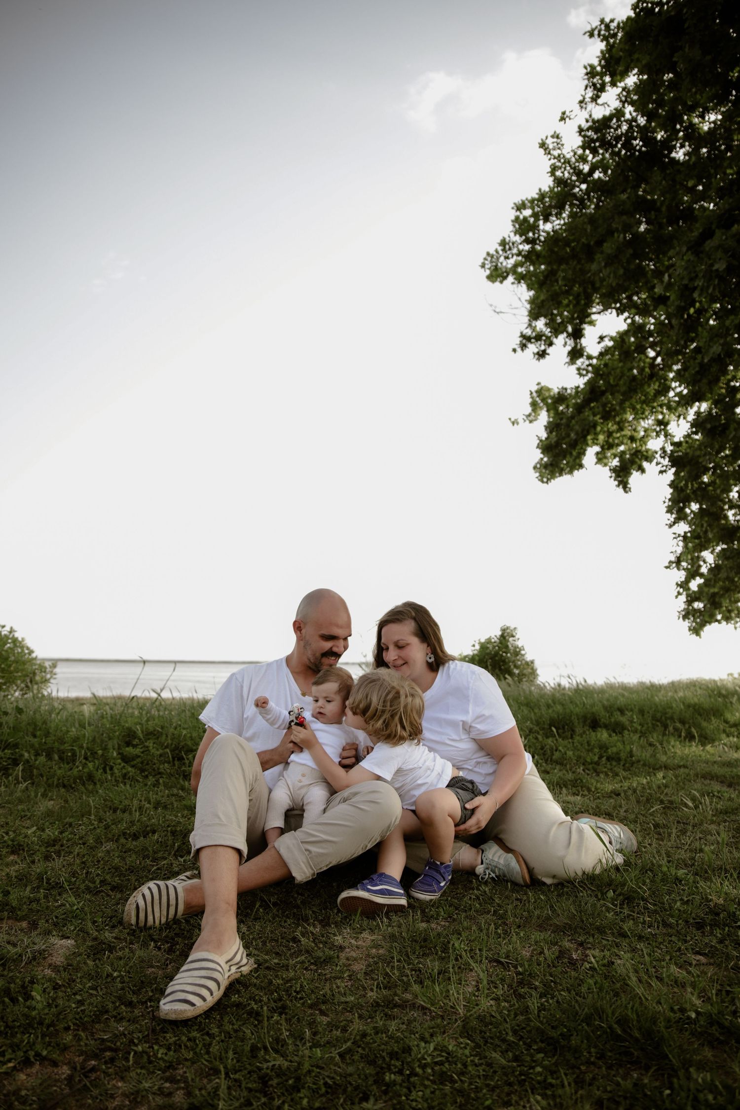 A family sits together on a grassy hillside, wearing matching white shirts and khaki pants on a cloudy day.