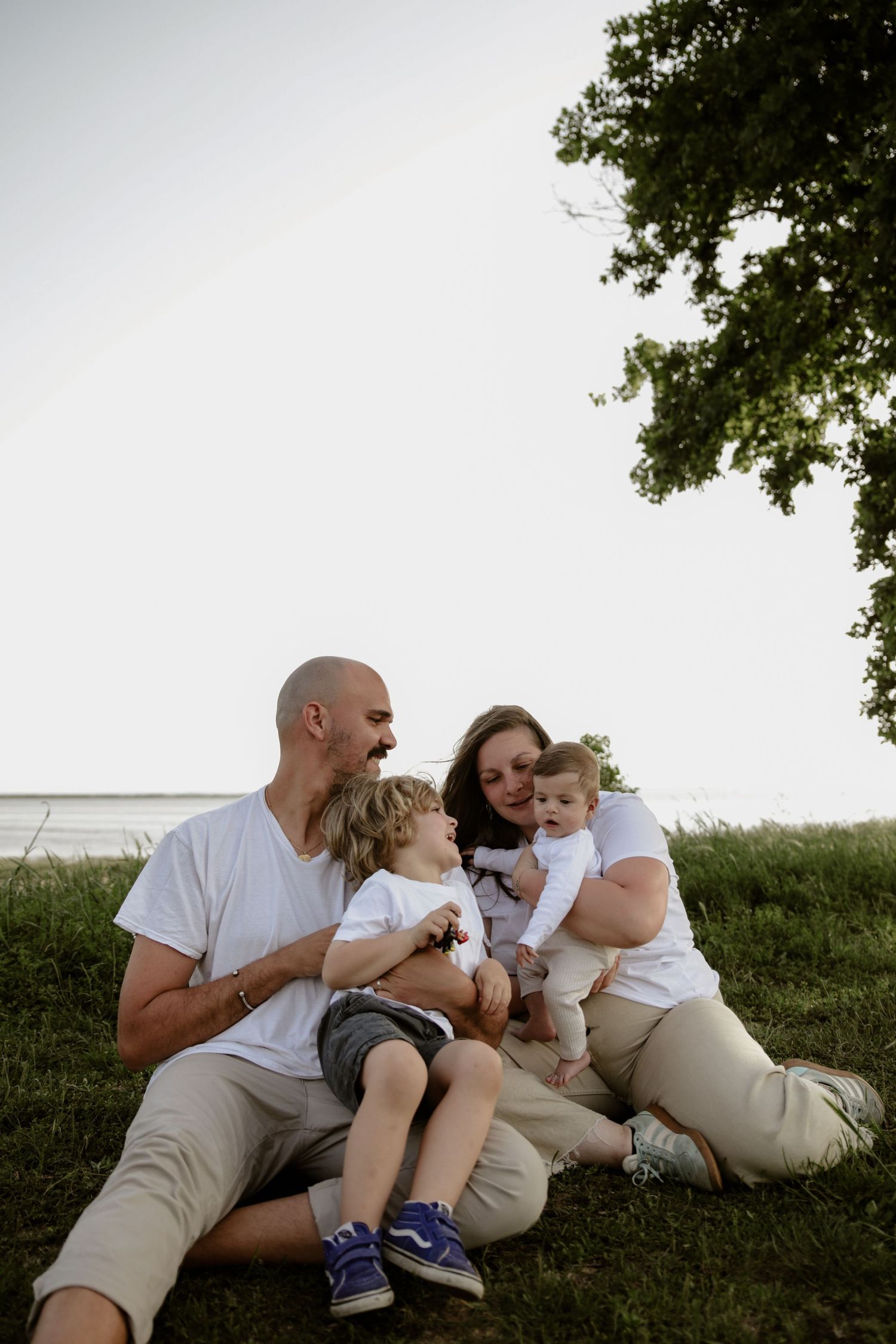 A family sits together on grassy hill overlooking water, wearing casual white and beige clothing.