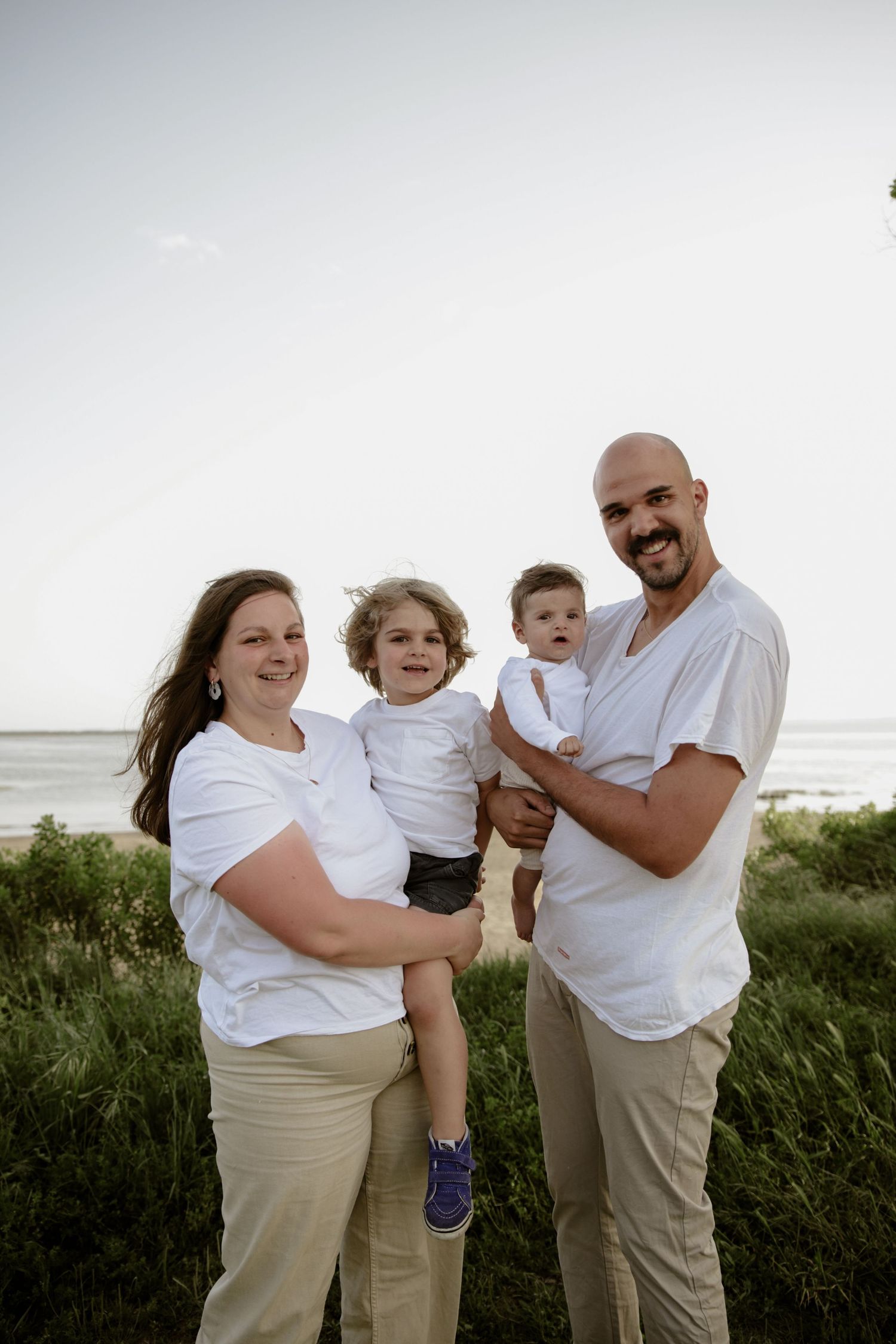 Family of four wearing white shirts and khaki pants pose together on grassy beach dunes at sunset.