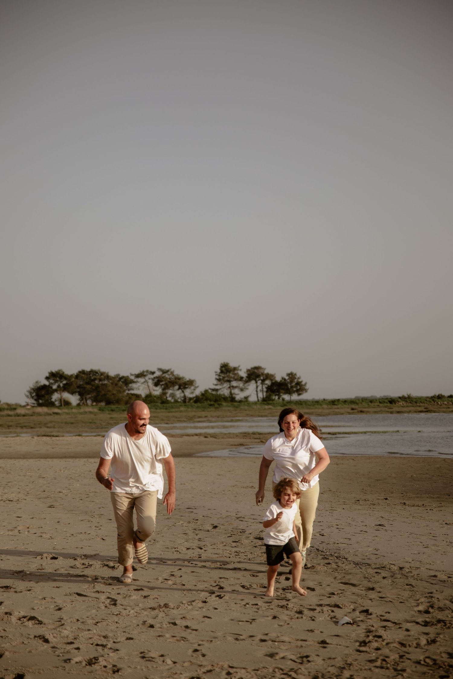 A couple playfully runs with their toddler on an empty beach at dusk.