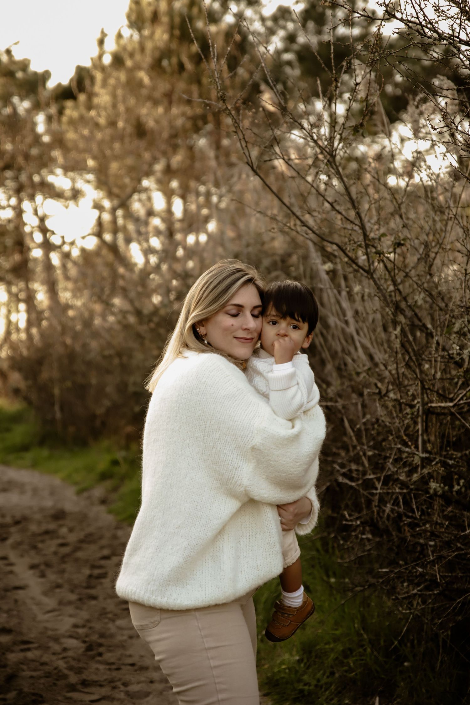 Mother holding child in cozy white sweater against sunset forest backdrop.