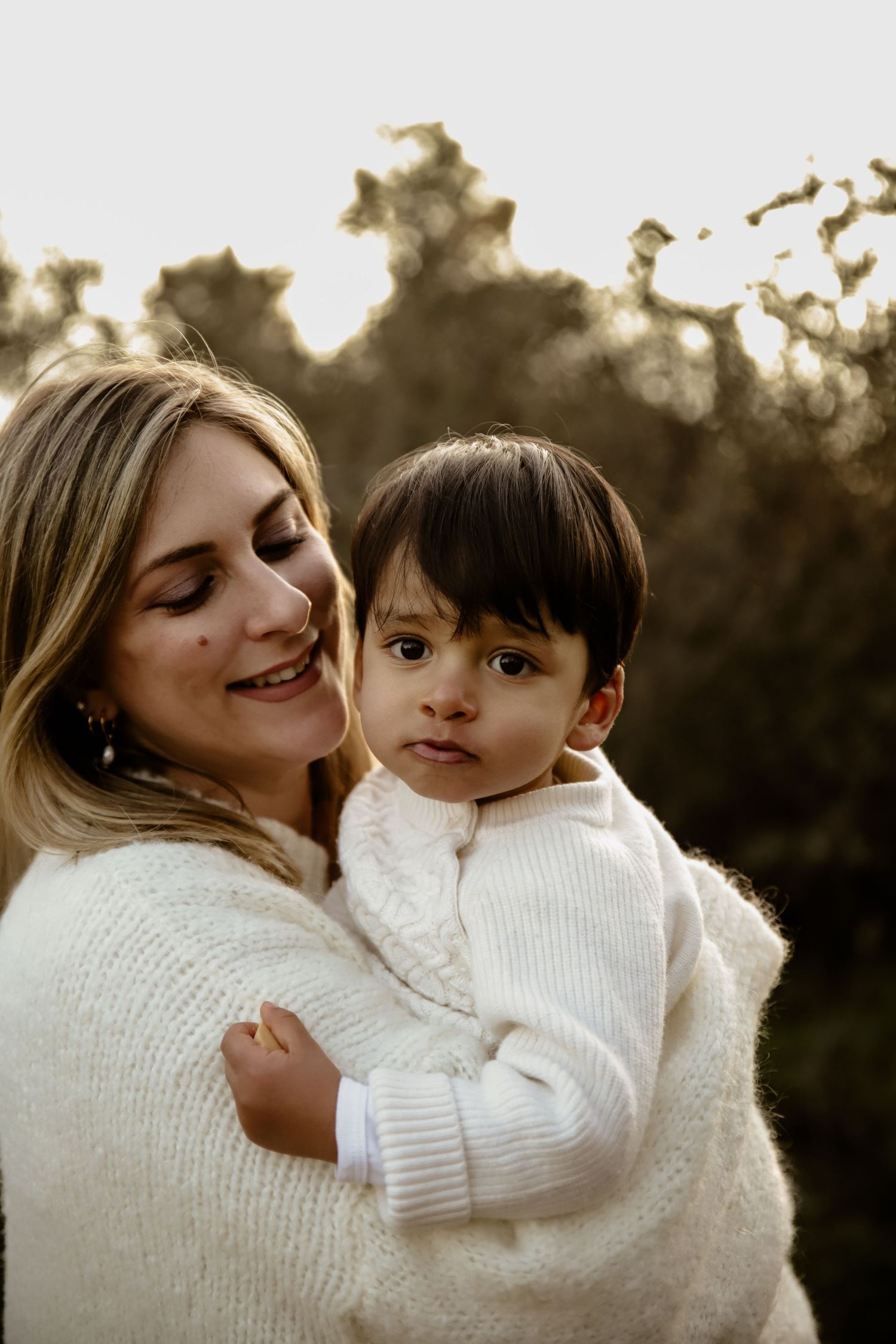 Mother and young child sharing tender moment in white sweaters at sunset.