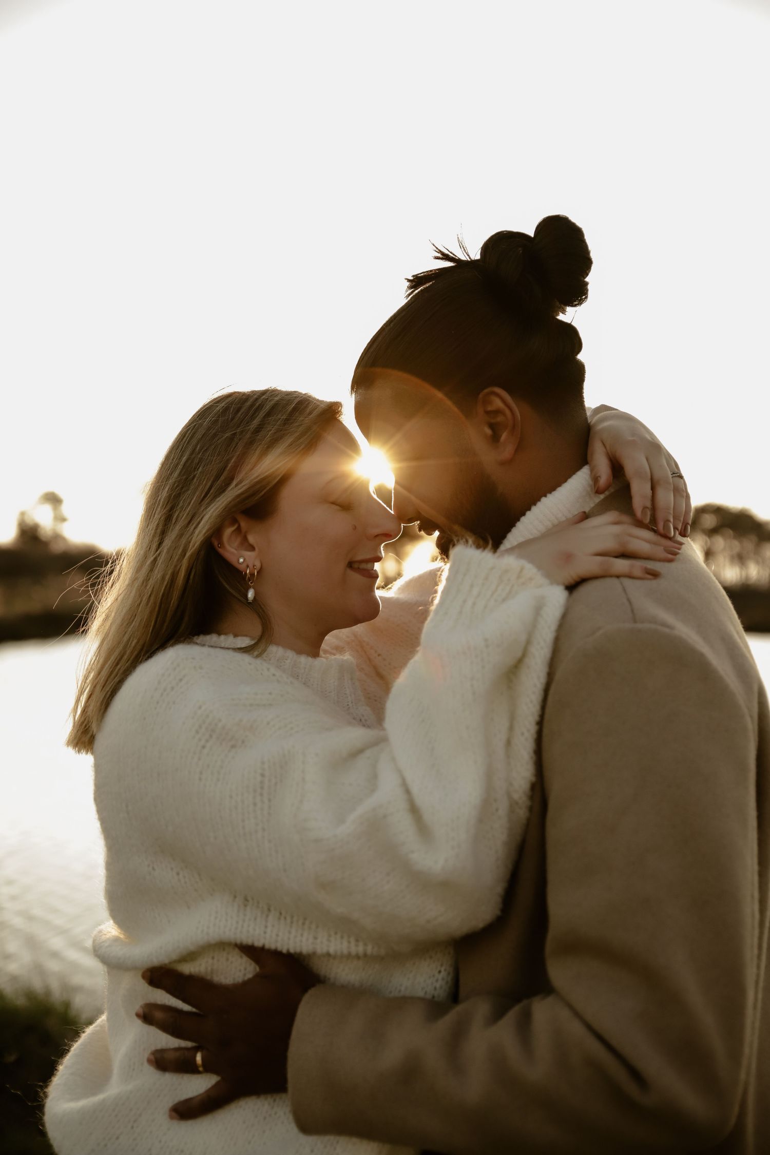 Couple in white sweaters sharing intimate moment at sunset by water.