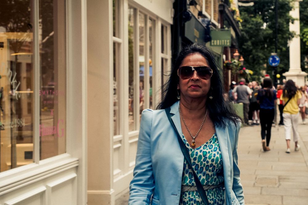 Woman in blue blazer and sunglasses walking past storefronts on a city street.