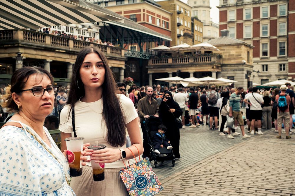 Two people holding drinks stand near a crowded cobblestone plaza in a historic city area.