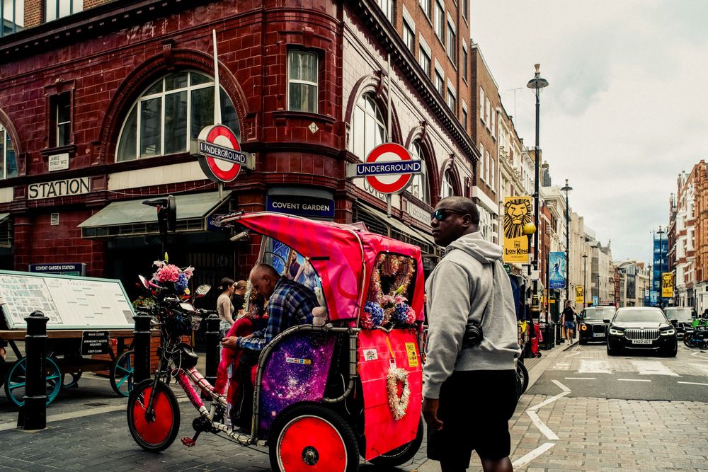 Red rickshaw or pedicab parked on London street near Underground station signs.