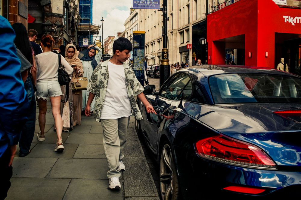 Busy urban street scene with pedestrians walking past parked cars.