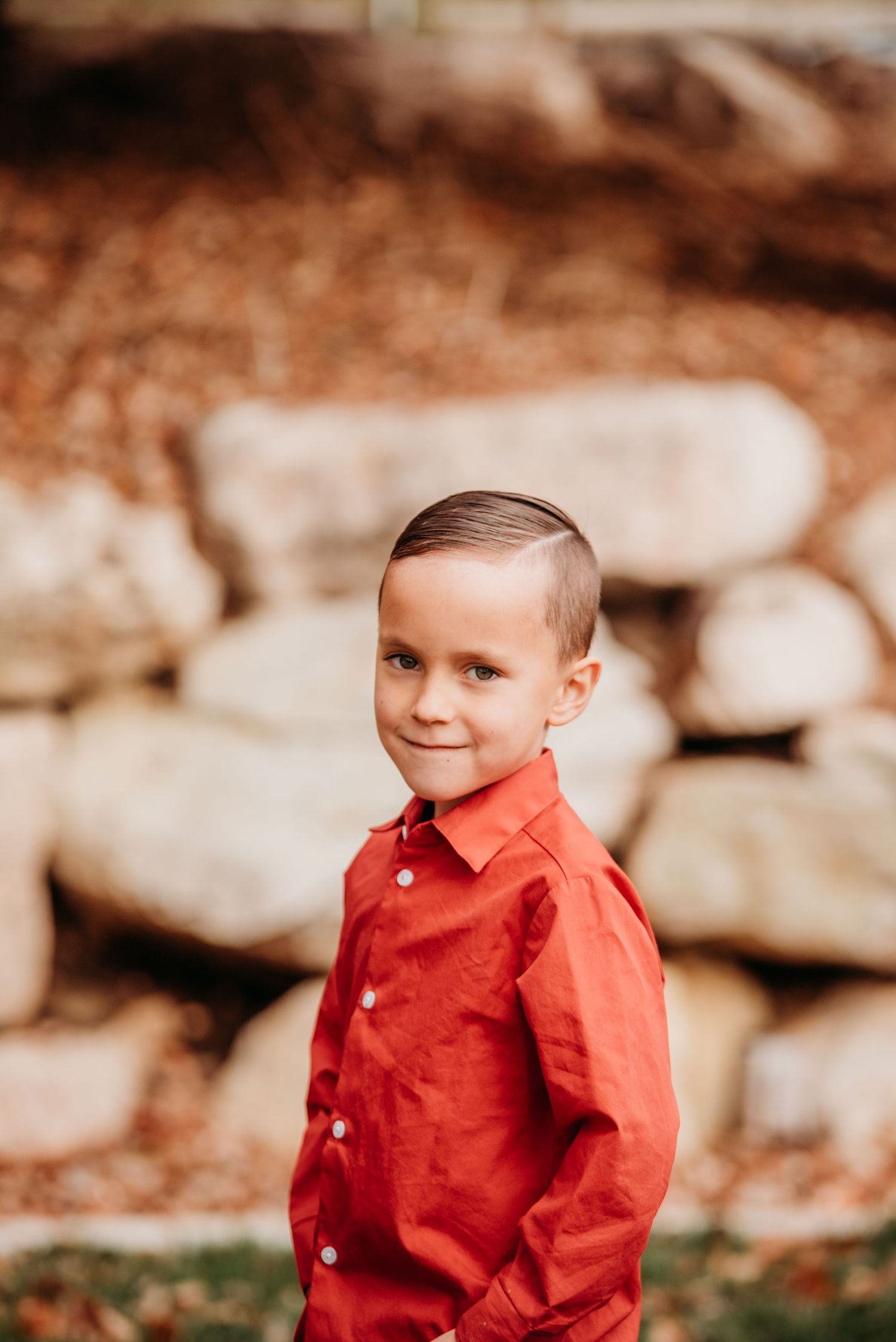 Young boy in a bright red button-down shirt smiling at camera in front of stone wall.