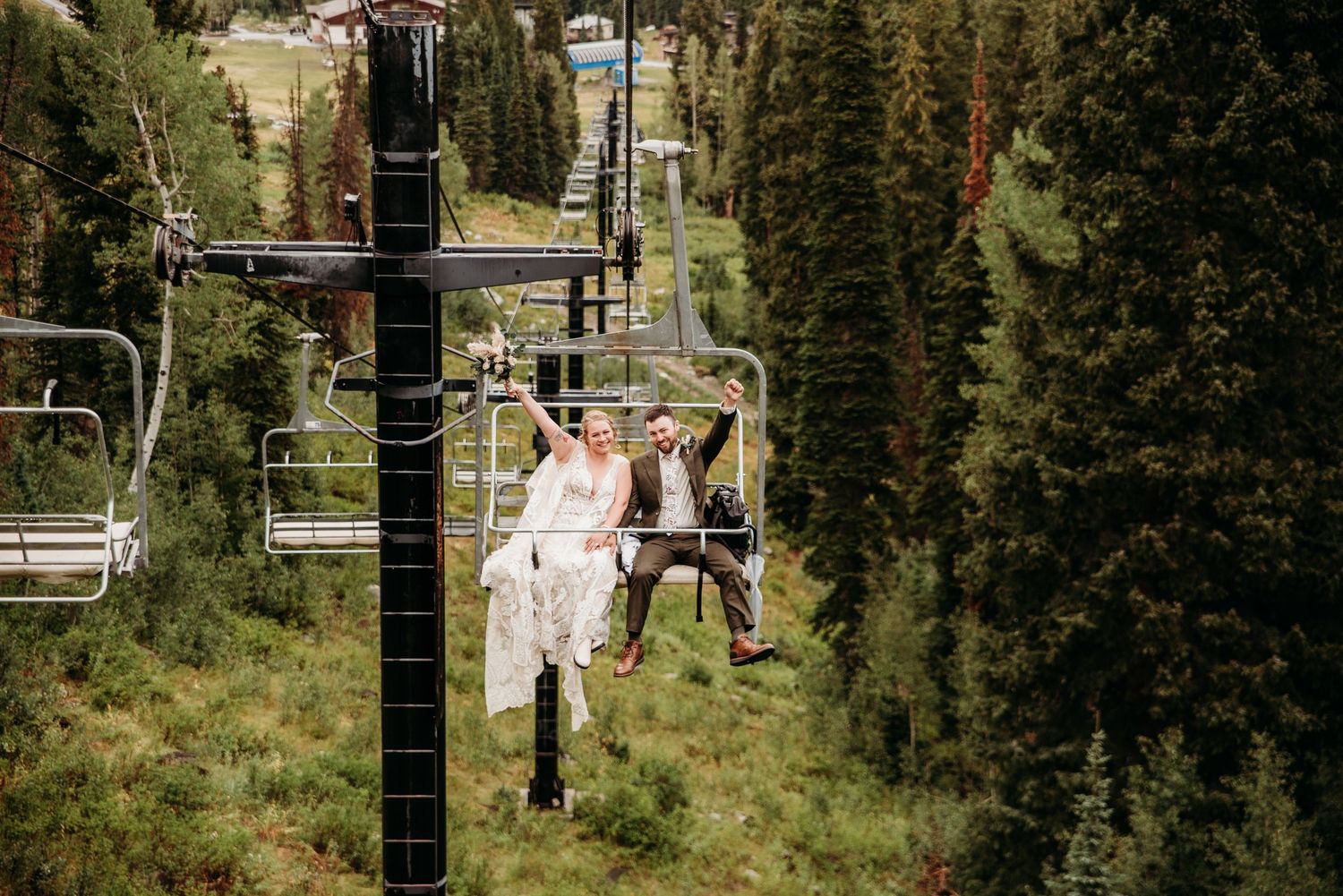 Wedding couple rides ski lift together in formal attire through mountain scenery.
