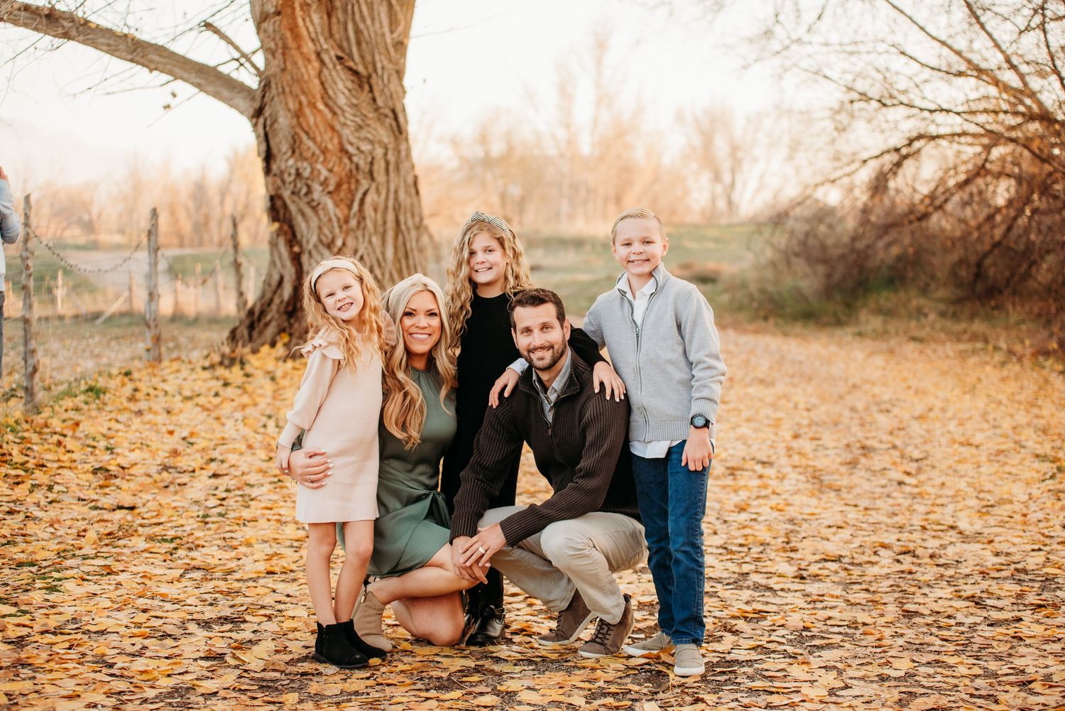 Family of five posing together on fallen autumn leaves under a large tree.