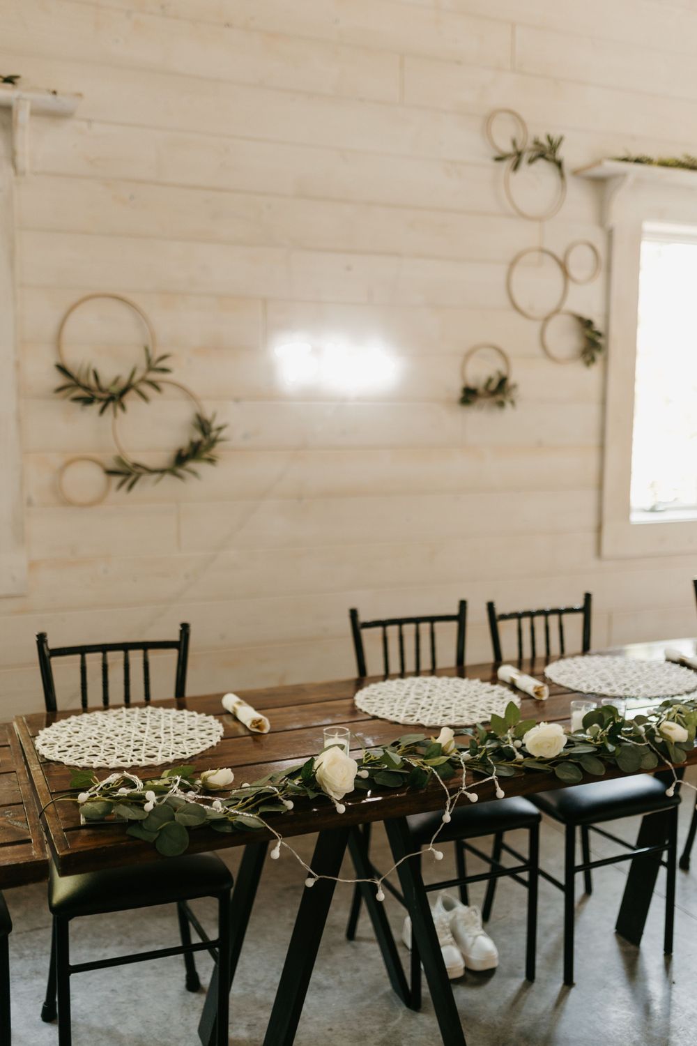 Rustic wedding reception table with greenery runner, hoop wall decor, and fairy lights in a barn.