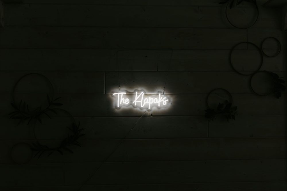 Glowing neon sign reading The Kleptics mounted on a wood-paneled wall with hoop greenery decor.
