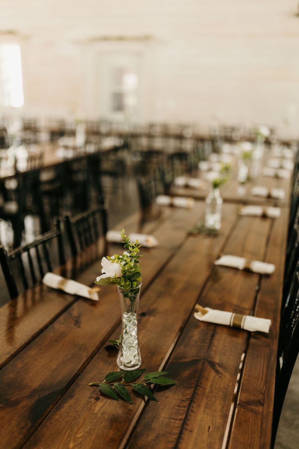 Long wooden farm tables set with bud vases, white flowers, and rolled napkins in a bright barn.