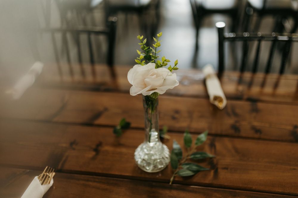 Single white rose in a crystal bud vase on a rustic wooden wedding reception table close-up.