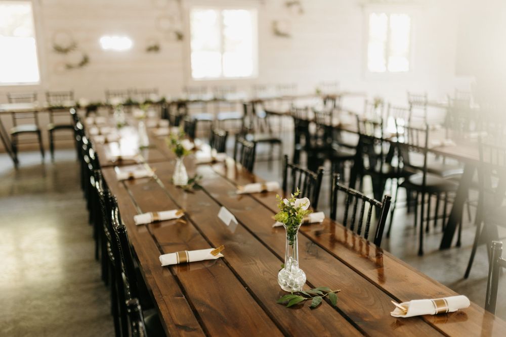 Bright barn wedding reception with long wooden tables, black chairs, and simple floral bud vases.