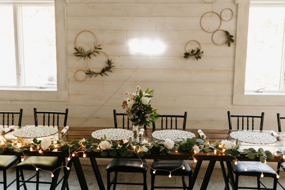 Elegant wedding reception table with greenery garland, string lights, and gold hoop wall decor in a white shiplap venue.
