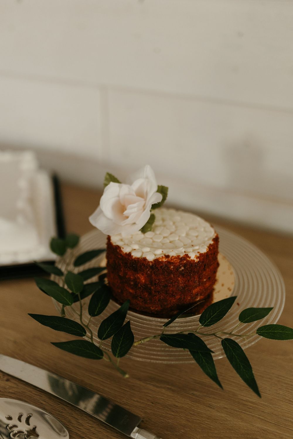 Close-up of a red velvet wedding cake with white frosting, pink rose topper, and greenery on a glass cake stand.