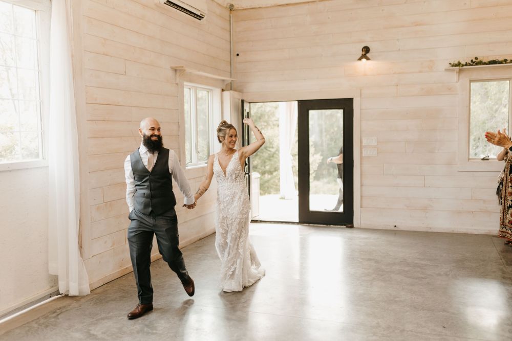 Bride in lace gown and groom in vest making grand entrance into bright white shiplap wedding reception venue.