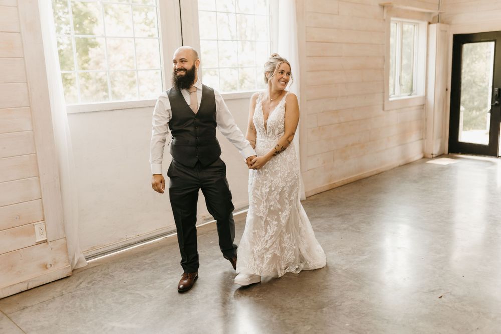 Bride and bearded groom walking hand in hand through a bright modern white shiplap wedding reception hall.