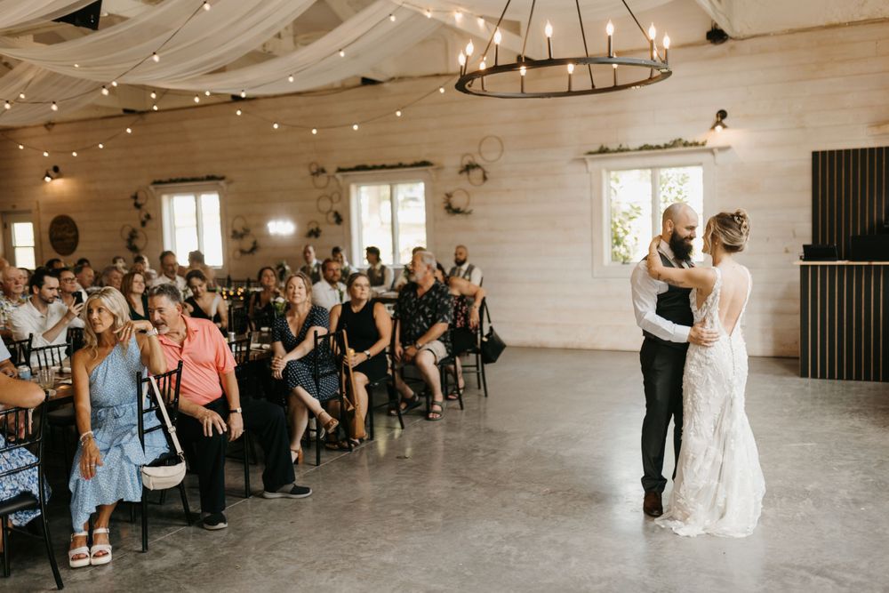 Bride and groom sharing first dance under chandelier with string lights while guests watch in rustic barn venue.