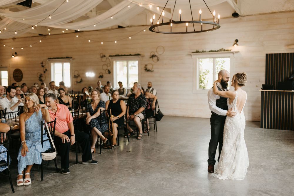 Newlyweds sharing an intimate first dance under a chandelier with string lights in a rustic white barn venue.