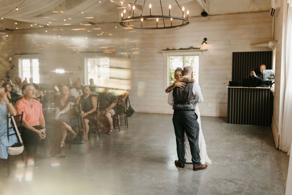Bride and groom embracing during first dance in a warmly lit rustic barn reception hall with seated guests.