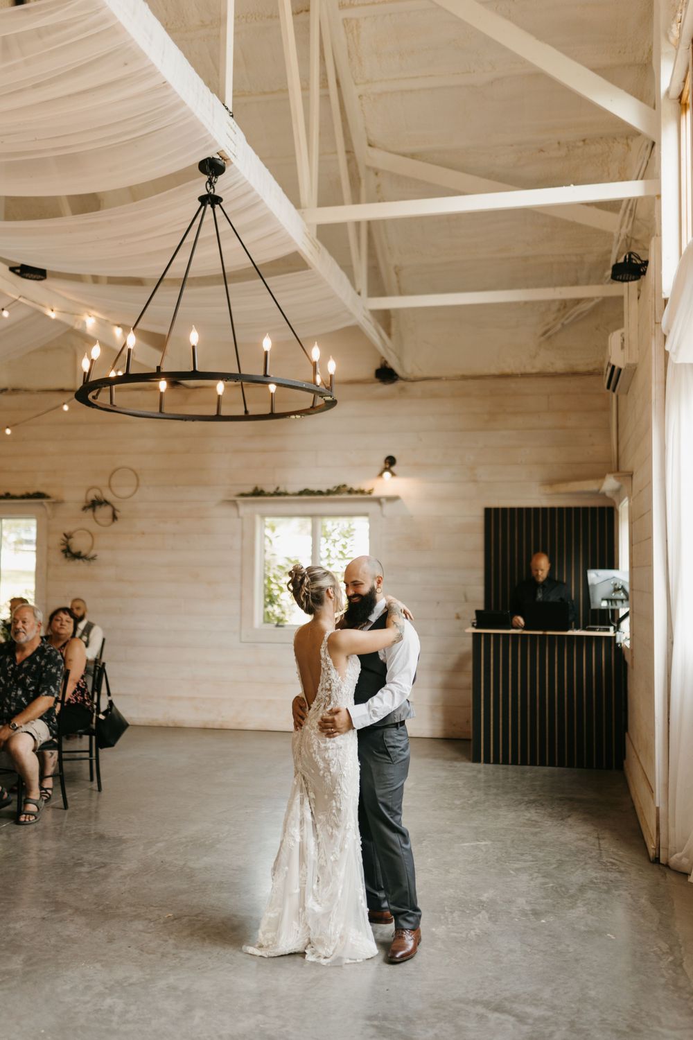 Bride and groom sharing first dance under a wrought iron chandelier in a bright white shiplap barn venue.