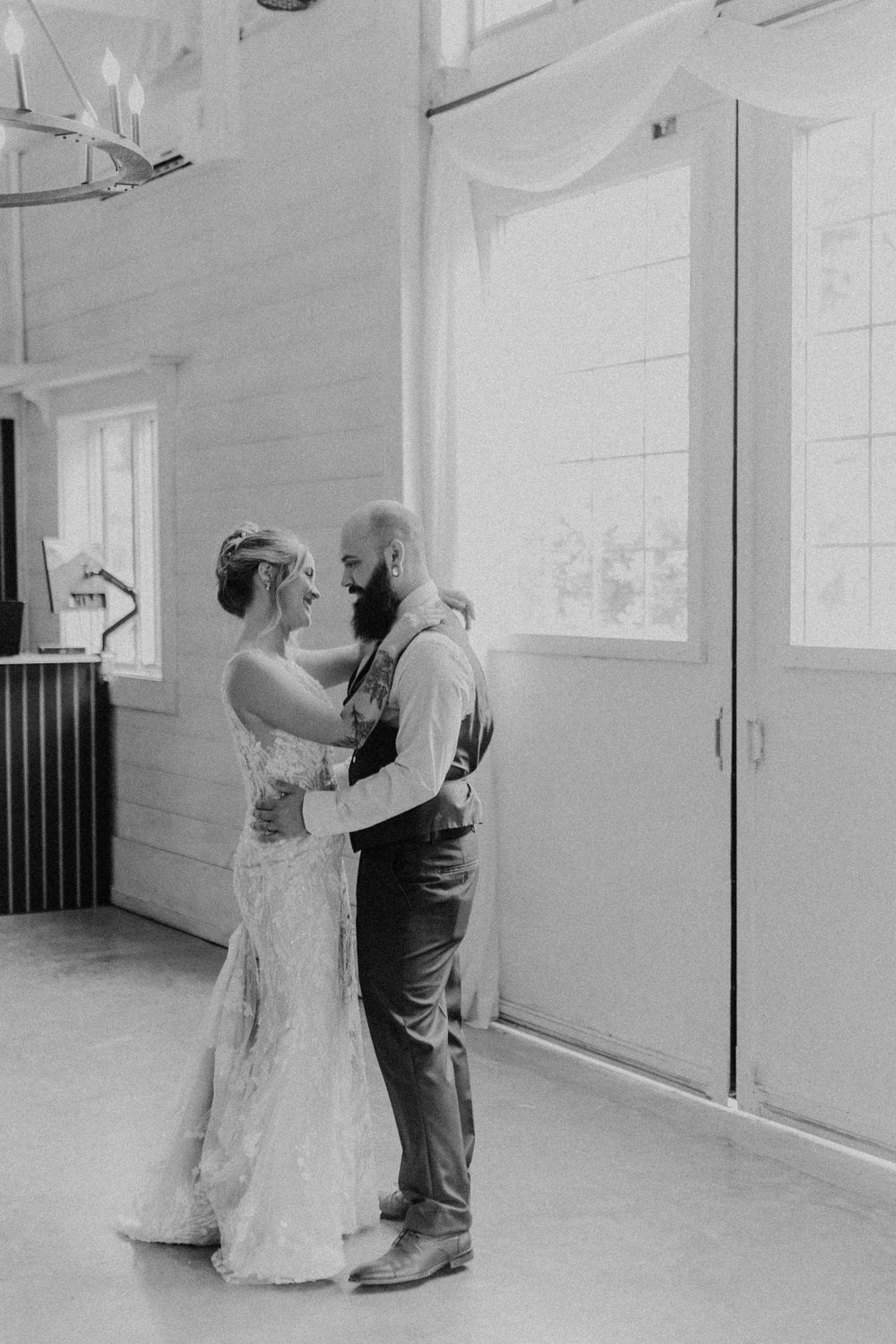 Black and white photo of bride and bearded groom sharing an intimate first dance in a bright barn venue.
