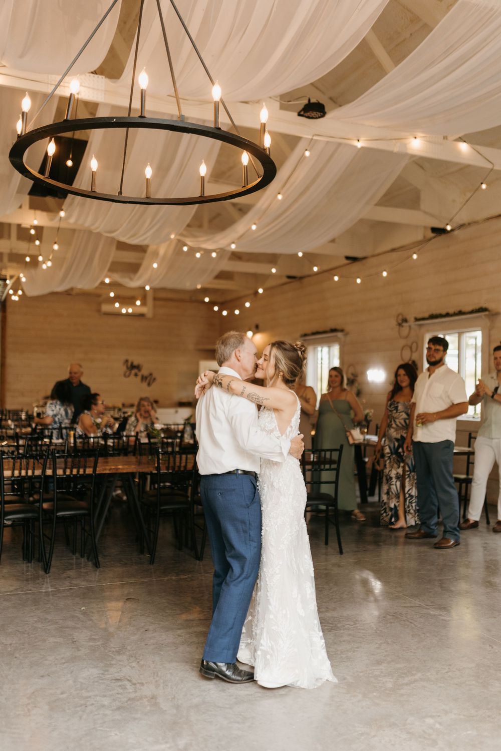 Bride and father share an emotional first dance in a rustic barn wedding venue with string lights.