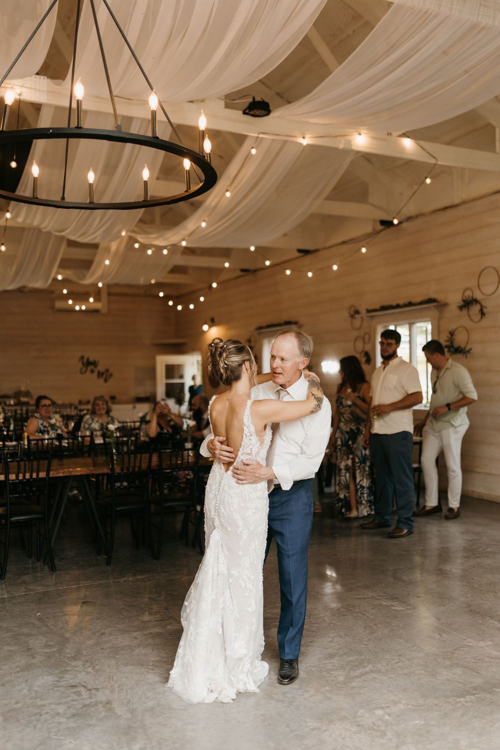 Bride in lace gown dances with father under warm string lights in a decorated barn reception hall.