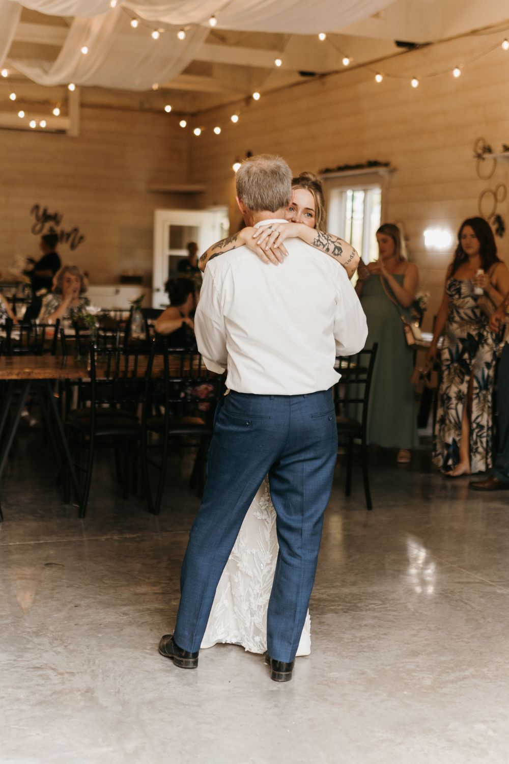 Father-daughter wedding dance captured from behind in a warmly lit rustic barn reception venue.