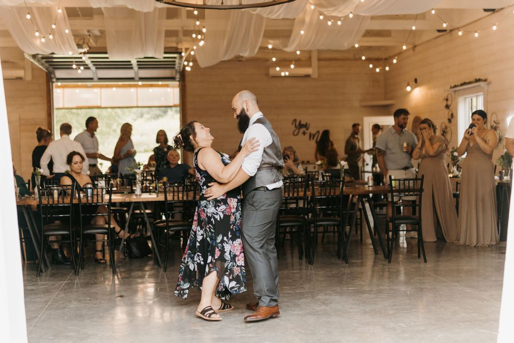Groom dances with mother in floral dress during wedding reception in a rustic barn venue.