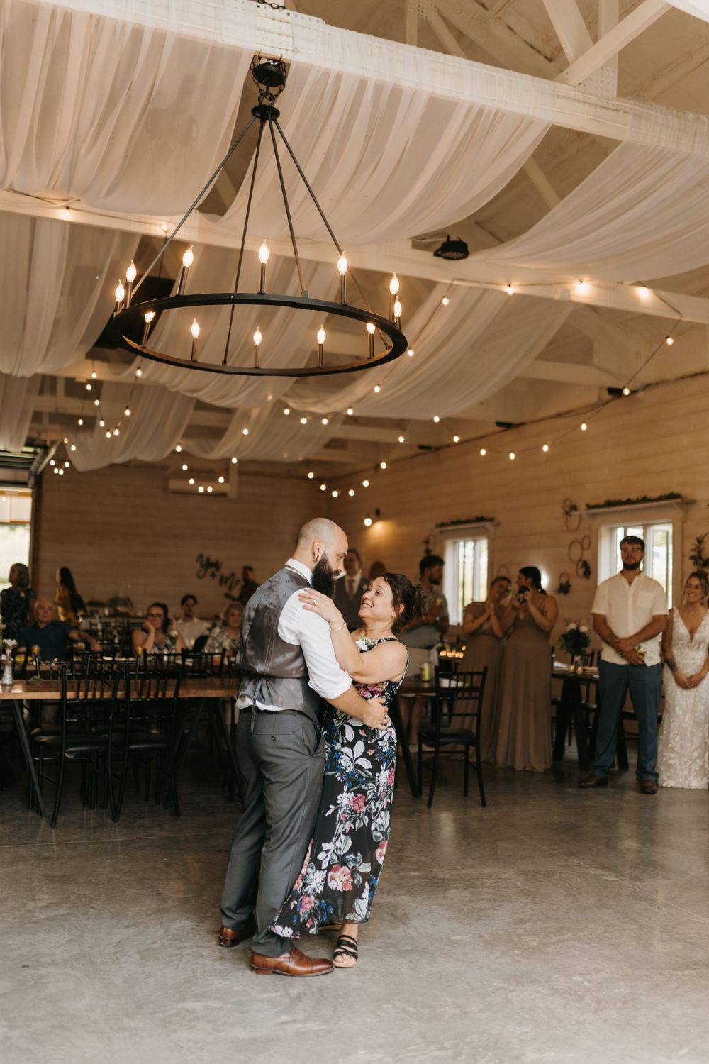 Groom and mother share a touching dance under a chandelier in a draped barn wedding reception.