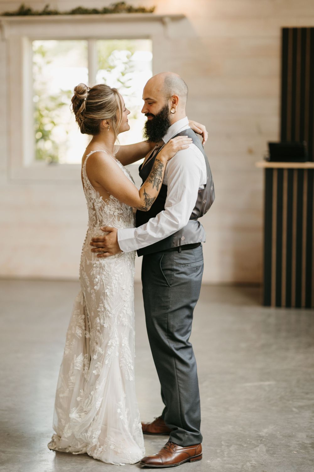 Bride and tattooed groom share an intimate first dance moment in a sunlit barn wedding venue.