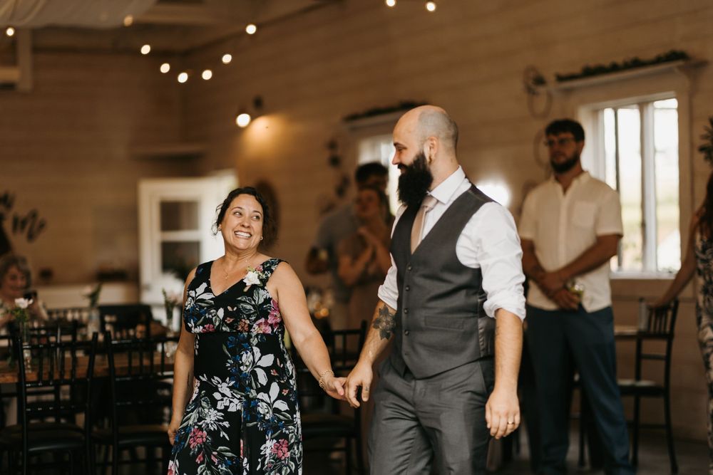 Smiling groom escorts mother in floral dress onto the dance floor at a rustic barn wedding.