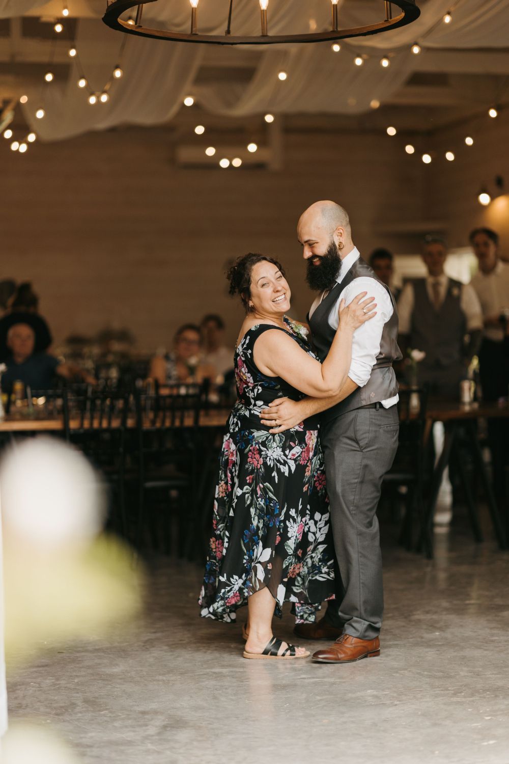 Groom dances joyfully with smiling mother in floral dress under string lights at wedding reception.