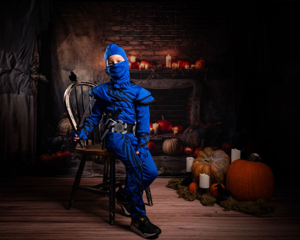 Blue ninja costume posed in wooden chair with Halloween decor and lit fireplace in background.