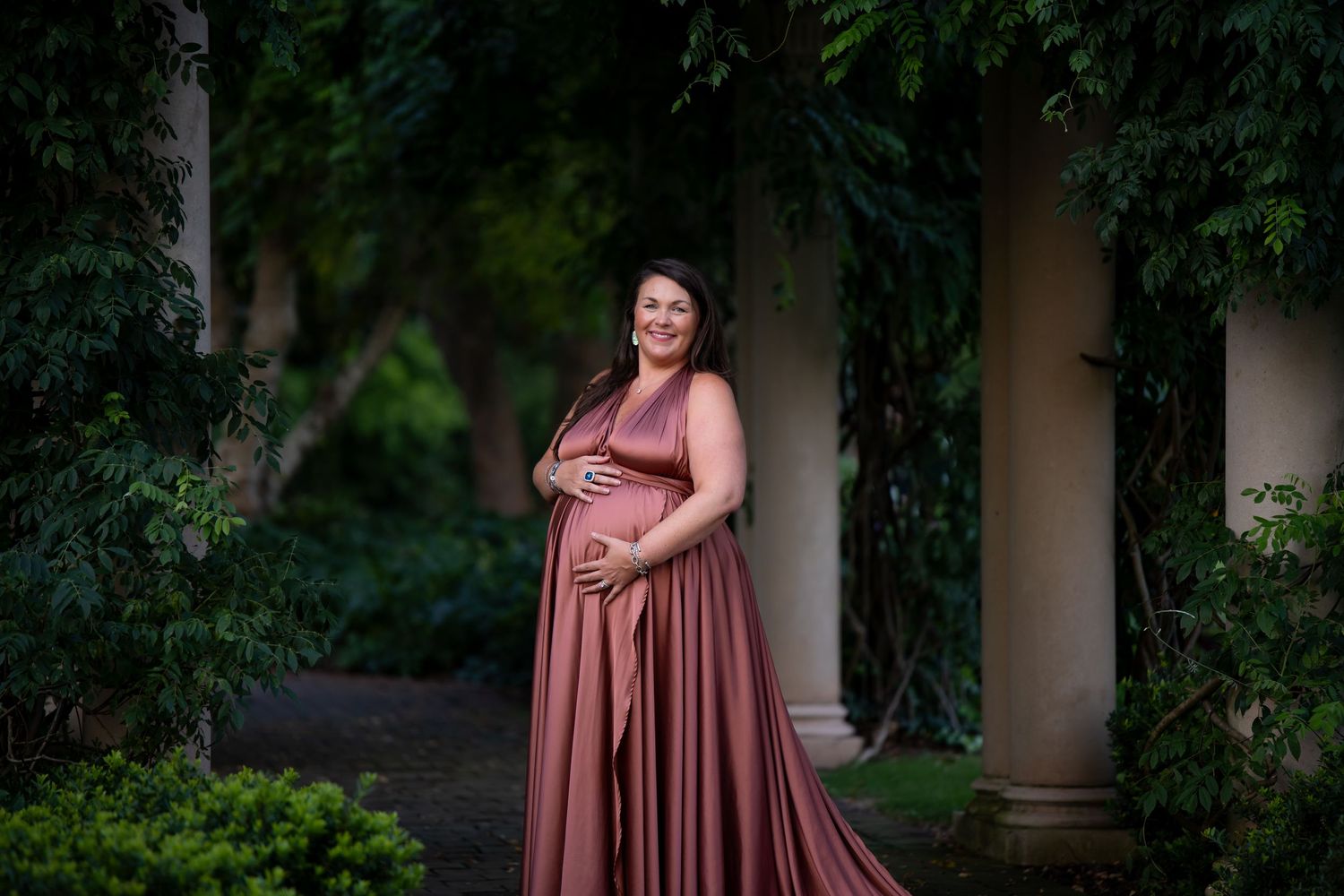 Maternity portrait of woman in flowing mauve dress standing among garden pillars.