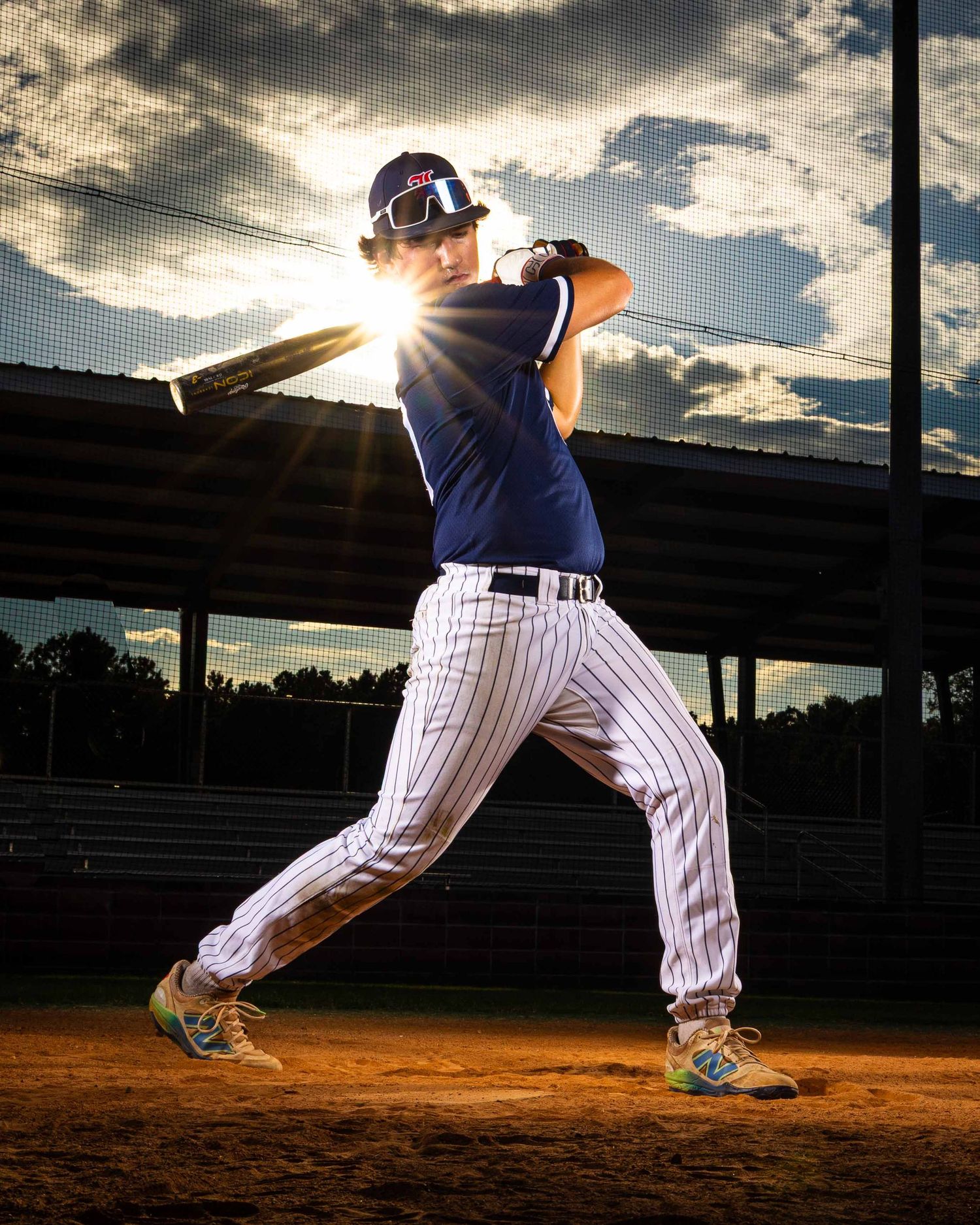 Baseball player in pinstripe uniform pitches at sunset with dramatic clouds and stadium silhouette in background.