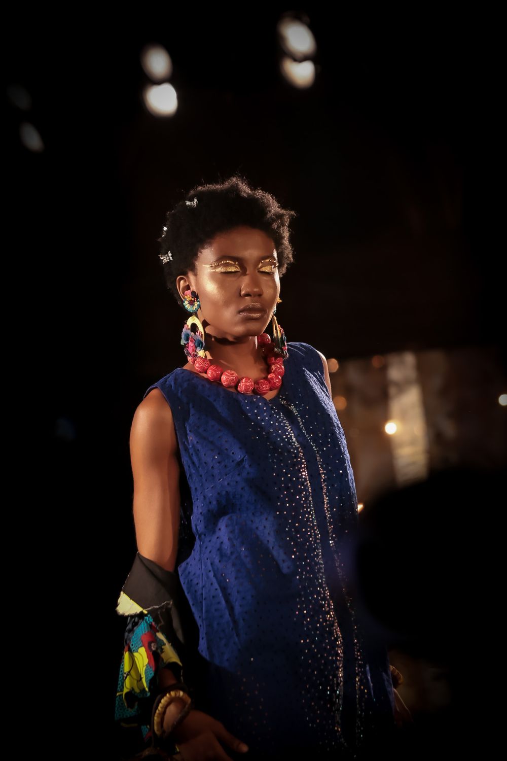 Model in sparkly blue dress and red necklace photographed during fashion show.